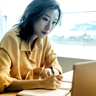 Photo of women writing in notebook while looking at laptop